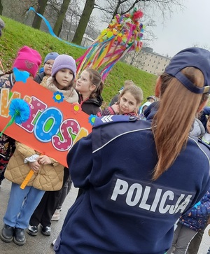 dzieci trzymające szyld z napisem wiosna, policjantka stojąca tyłem do obiektywu aparatu